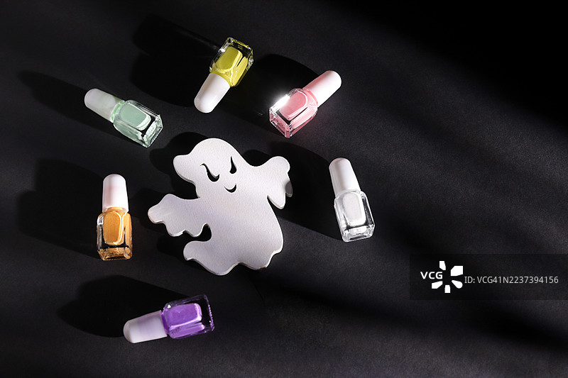 从高处俯瞰一个白色的幽灵装饰，周围环绕着彩色的指甲油瓶，背景为深色，非常适合万圣节美容主题。图片素材