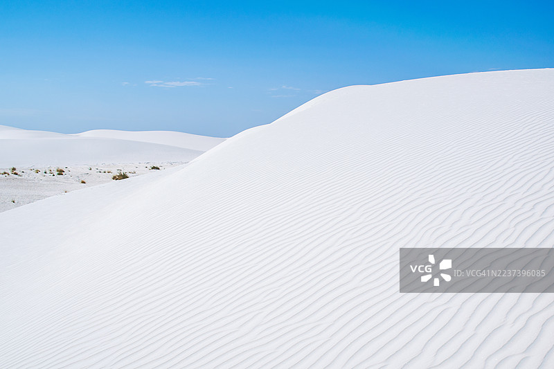 在晴朗的蓝天下，白色的沙丘呈现出壮丽的景色，展示了自然的纹理和图案。图片素材