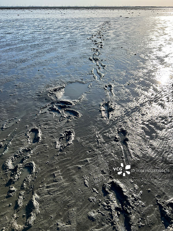 高角度拍摄的退潮时海滩湿泥沙上的脚印，远处是明亮天空下的风力涡轮机，地点：兰格奥格，德国图片素材