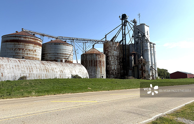 从低角度拍摄的生锈的谷物升降机和储仓，背景是晴朗的蓝天，旁边是一条乡村公路。图片素材