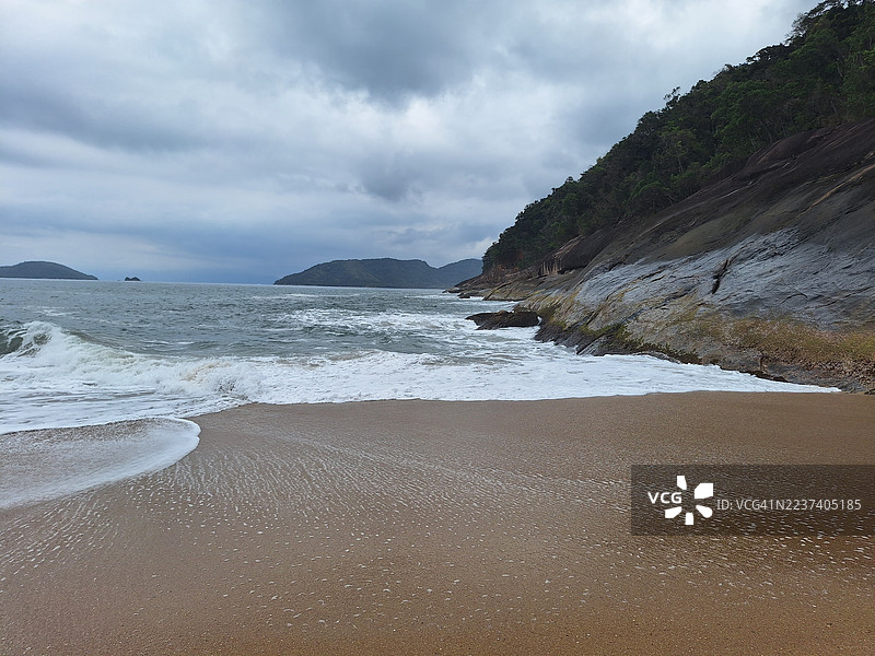 热带海滩的风景，海浪拍打着海岸，多云天空下的茂密绿色山坡。图片素材