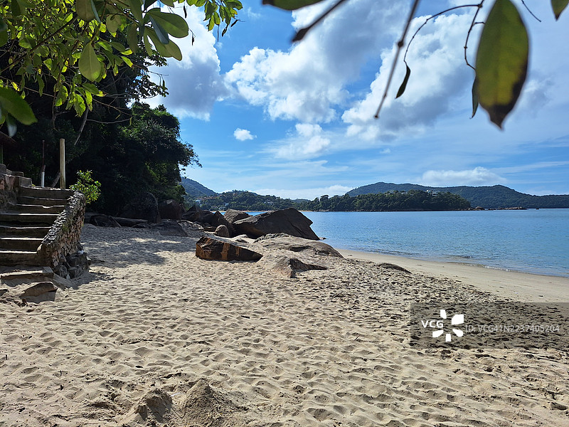 宁静的沙滩，郁郁葱葱的绿叶和清澈的蓝水，在明亮的天空下，提供了一个宁静的避世之所。图片素材