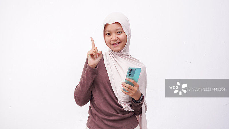 戴着头巾的快乐女孩，手持智能手机，对着镜头微笑，手指指向上方，白色背景上的人物肖像图片素材