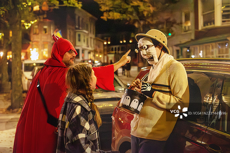 53岁的爸爸穿着恶魔服装，顽皮地触碰着他19岁儿子戴的木乃伊面具上的眼球。他们10岁的女儿在旁边笑着，三人站在宾夕法尼亚州吉姆·索普的夜间街道上。图片素材