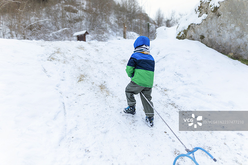 一个男孩在雪地里拉着他的雪橇上坡。图片素材