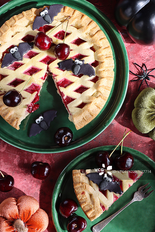 万圣节派对食物的图片，绿色盘子上摆放着新鲜出炉的樱桃挞，金色格子酥皮顶部装饰着黑色蝙蝠翻糖糖霜，切好的派块上有樱桃，旁边有一个毛绒南瓜，背景为斑驳的红色，俯视角度。图片素材