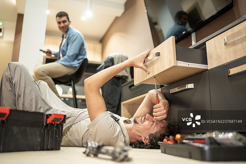 居家家具组装厨房柜下方失误：男子躺在地板上摆弄工具，朋友在一旁观看图片素材