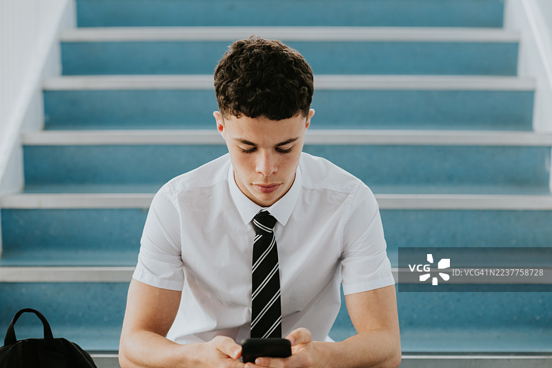 一名青少年男学生坐在学校的楼梯上，低头看着他的智能手机。留有文字空间。图片素材