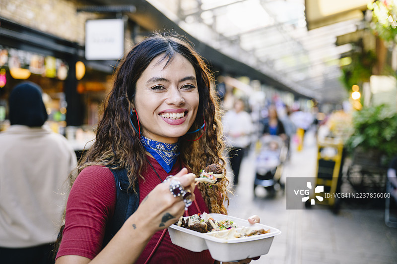 一位快乐的中年女性肖像，她正对着镜头微笑，并从外卖盒里吃沙拉三明治。图片素材