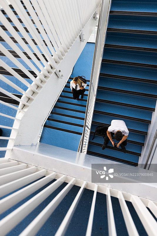 高角度拍摄：一名女高中生背着包走上楼梯，去见一个坐在楼梯上方、正看着智能手机的男高中生，地点在英国纽卡斯尔泰恩河畔。图片素材