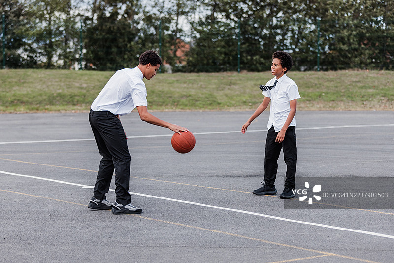 英国纽卡斯尔泰恩的户外硬地球场，两名十几岁的男孩在休息时间于中线处打一场一对一的篮球赛，摄于英国一所高中。图片素材