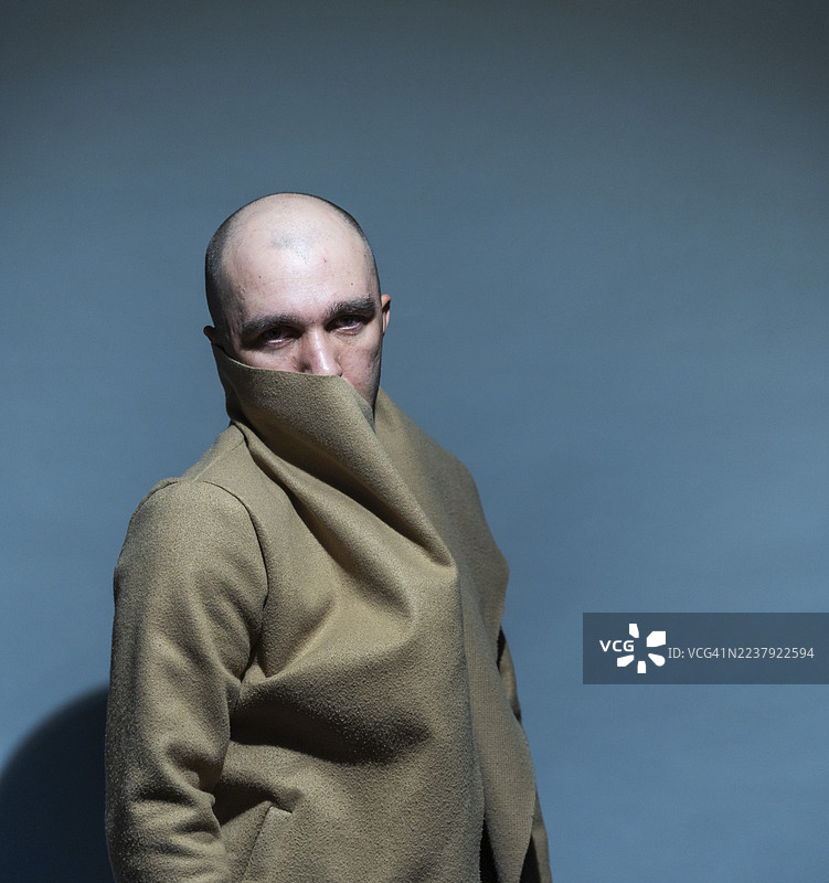一位秃顶男子用一件温暖时尚的外套遮住脸部下半部分，在工作室的蓝色简约背景下，展现神秘、匿名和内省的概念图片素材