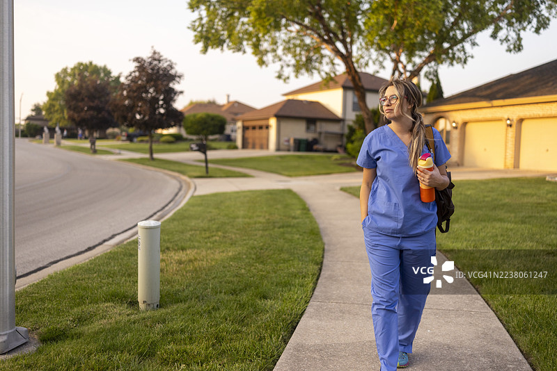 身穿蓝色制服的女护士在住宅区外走，手里拿着水瓶，要去上班。图片素材