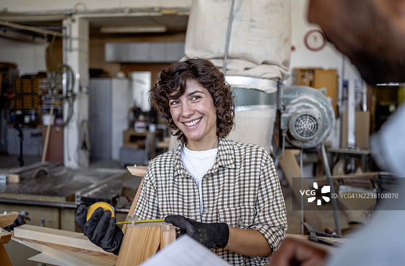 一位微笑着的女木工在繁忙的木工车间里拿着卷尺图片素材