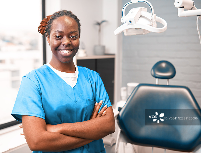 身穿蓝色手术服的牙科专业人士，面带微笑地站在现代牙科椅和设备旁图片素材
