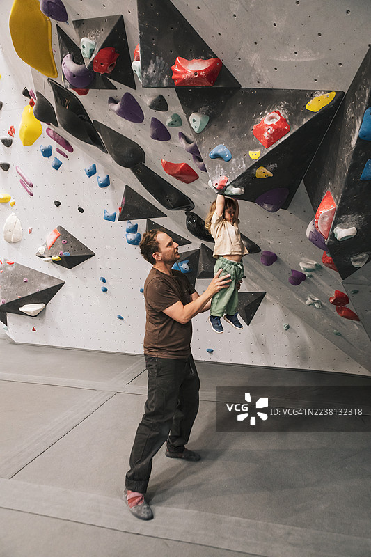 父亲在攀岩馆里，帮助他年幼的女儿抓住攀岩点。图片素材