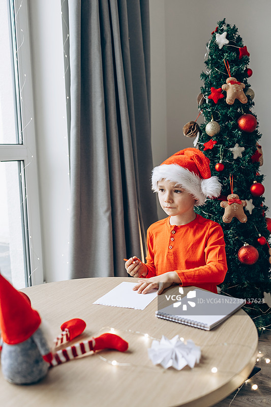 一个戴着圣诞帽的孩子在家给圣诞老人写信，坐在圣诞树旁。温馨的节日场景，装饰有节日气氛，自然光照射，捕捉了圣诞节和节日传统的精神。图片素材