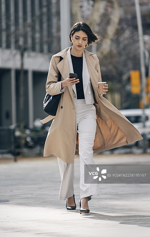 自信的女商人在户外穿着时尚的风衣，手里拿着咖啡。图片素材