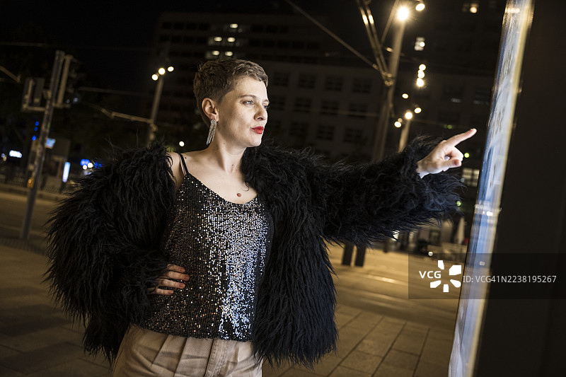 在夜晚的城市中，富有表现力的成年女性与数字信息板互动图片素材