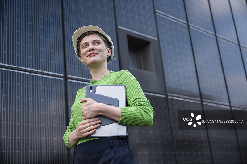 戴着头盔和手持平板的工程师在户外的太阳能面板墙旁站立图片素材