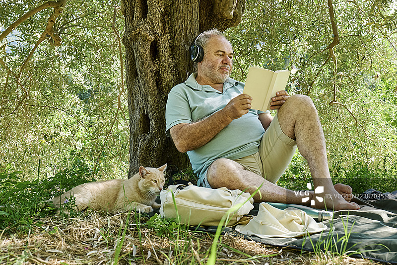 男人在树下放松，手里拿着书和耳机，旁边有一只猫。图片素材