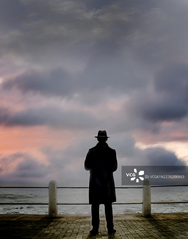 神秘的身影，穿着长外套和帽子，面朝海洋，夕阳下的景象图片素材