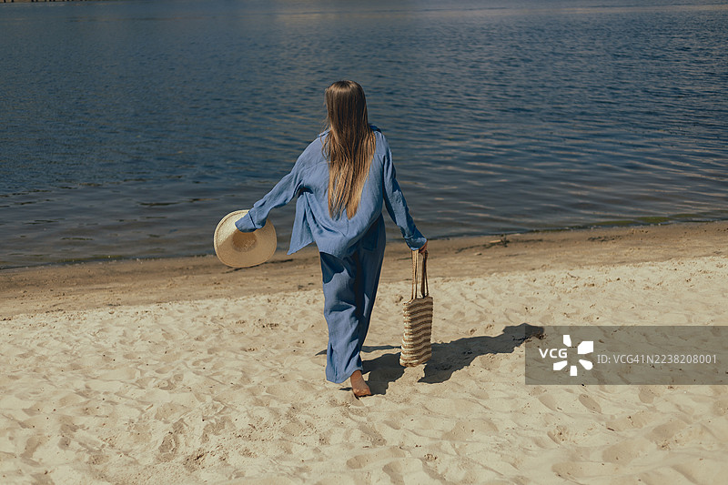 一位身穿蓝色衣服的年轻女子，一手拿着宽檐帽，一手拎着草编包，正走向水边，漫步在沙滩上。图片素材