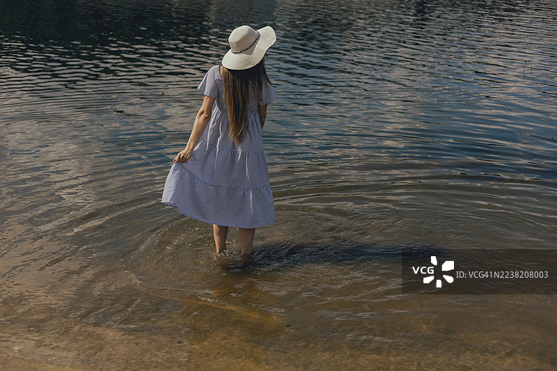 一位年轻女子身穿连衣裙，头戴宽檐帽，在夏日的海边站立。图片素材