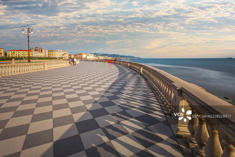 意大利利沃诺的马斯卡尼露台海滨长廊，在金色时刻，行人沿着标志性的棋盘格露台漫步，旁边是平静的第勒尼安海和色彩缤纷的建筑。图片素材