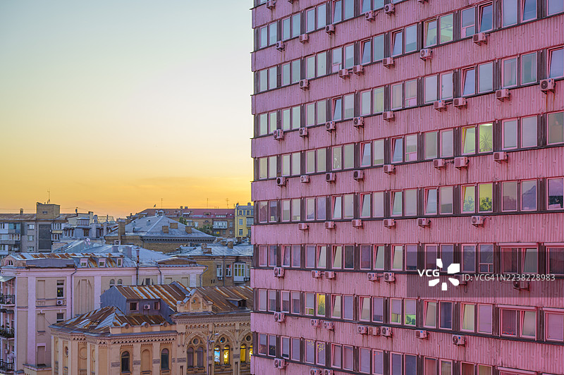 粉色公寓楼，有成排的窗户和外部空调，背景是温暖的日落天空，在密集的城市景观中形成新旧城市立面的对比。图片素材