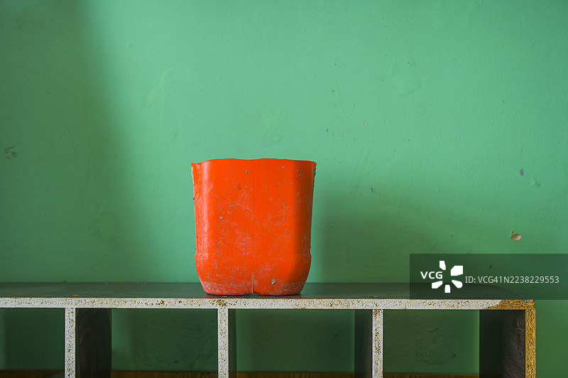 橙色水桶盆放在一个风化、简约的金属架上，在抽象静物构图中与鲜艳的绿墙形成鲜明的色彩对比。图片素材