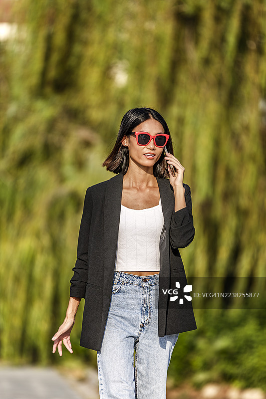 一名身穿黑色西装外套、戴着红色太阳镜的女子在户外边走边打电话。西班牙图片素材