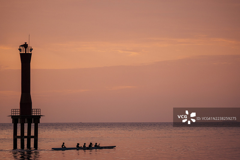 日落时分，剪影中的人们在宁静的水面上划着独木舟，旁边有一座灯塔。地点：印度尼西亚西苏门答腊省巴东。图片素材