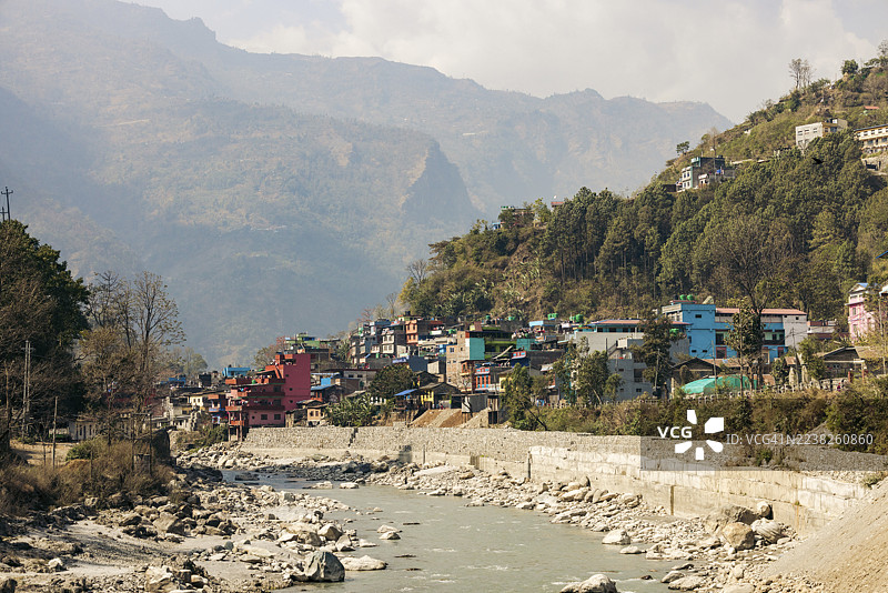 部分多云天气下，色彩缤纷的建筑沿河岸排列，背景是连绵的山峦。位于尼泊尔甘达基省穆斯丁县的安纳布尔纳。图片素材
