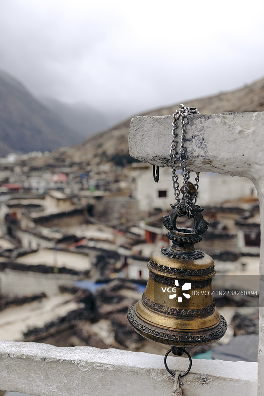 背景为模糊的山村和朦胧的天空，栏杆上放着一个黄铜铃铛。尼泊尔甘达基省马斯塘县安纳普尔那地区马尔法。图片素材