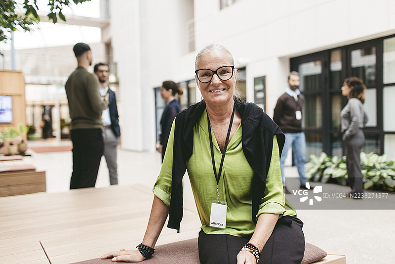 一位戴眼镜、面带微笑、自信的女性商务专业人士坐在会展中心的板凳上。图片素材