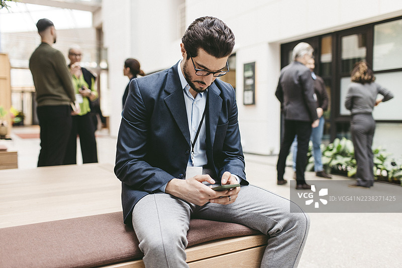 专注的男性商务人士在会展中心大堂的长椅上使用智能手机。图片素材