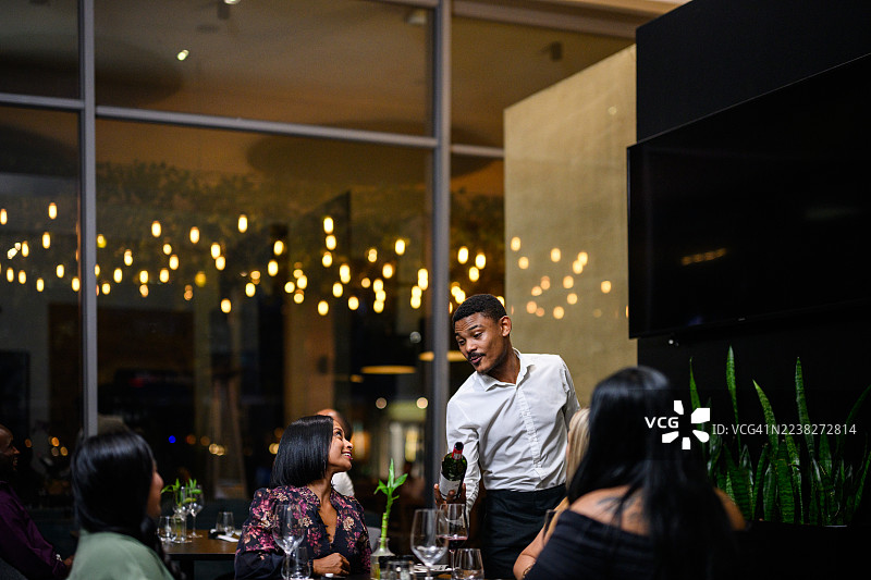 侍者在奢华餐厅内为一同用餐的女性朋友们奉上葡萄酒，她们身后是闪耀的金色灯光。图片素材