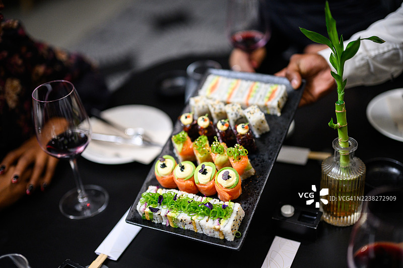 餐厅餐桌上摆放着寿司拼盘、微型蔬菜、三文鱼玫瑰、时尚三明治和加州卷，近景拍摄图片素材