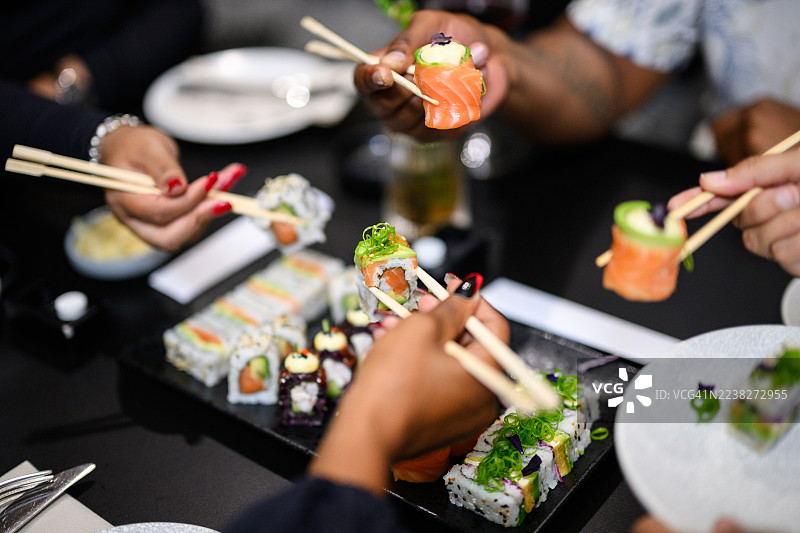 在餐厅的桌子上，人们从共享的寿司拼盘中取用三文鱼玫瑰寿司和加州卷，拼盘上装饰有微型蔬菜图片素材