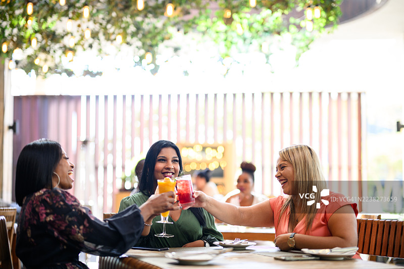 在有顶灯的绿叶户外餐厅露台上，快乐的女朋友们举杯畅饮彩色饮品图片素材