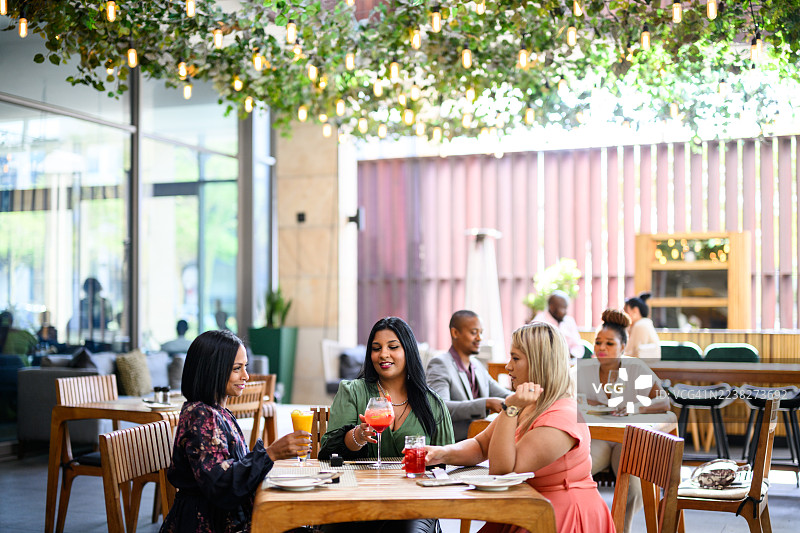 明亮露台餐厅的广角镜头，三位不同肤色的女士在头顶灯光下，一边聊天一边拿着色彩缤纷的饮品图片素材