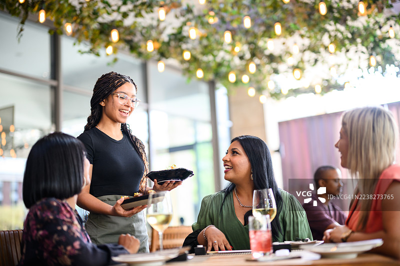 一群女性在一家明亮露台餐厅里交谈，服务员展示着食物，餐厅装饰有绿叶和吊灯。图片素材