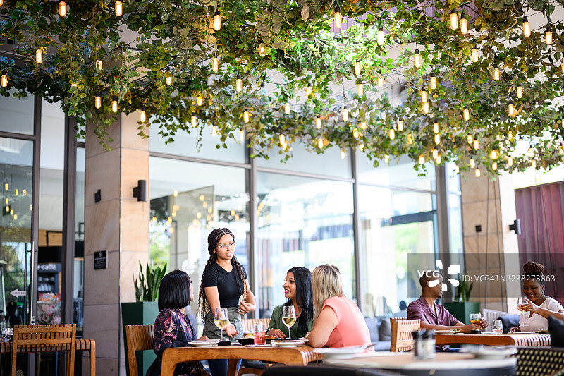 宽幅视角，服务员在绿植丰富、悬挂着灯饰的明亮露台餐厅里，为一群女性顾客提供服务图片素材