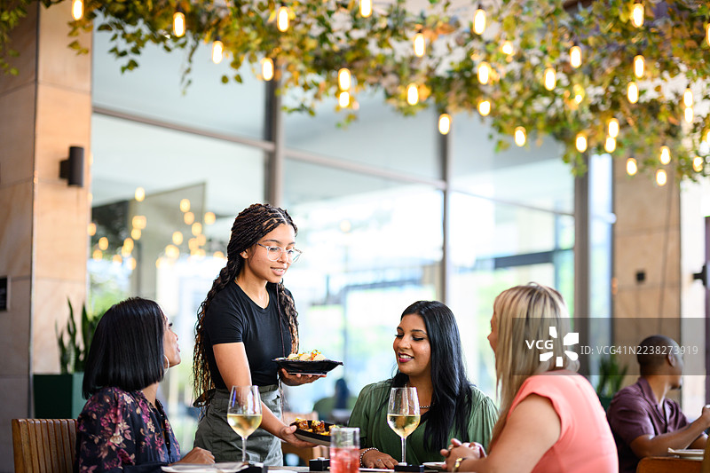 服务员在绿植和暖色灯光下的露台餐厅为一群用餐的女士们上菜图片素材