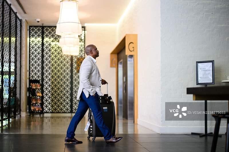 商务男士侧身拉着行李箱，走过拥有现代室内装饰和吊灯的现代酒店大堂图片素材