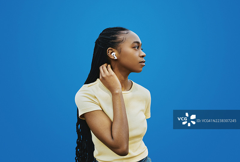 穿着黄色衬衫的非洲裔美国女性戴着耳机，在蓝色背景前若有所思地听音乐图片素材