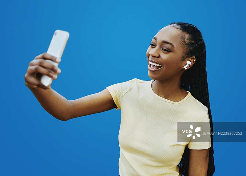 快乐的非裔美国女性，戴着无线耳机，在蓝色背景前微笑自拍。图片素材