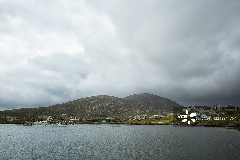外赫布里底群岛巴拉村的景色，山坡上有古老的石头房屋，低沉的深灰色云层，以及一个停泊着船只的小型码头。图片素材