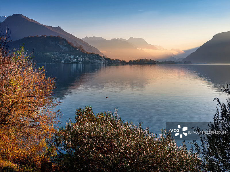 瑞士卢加诺，秋日宁静湖泊映照着雄伟的山峦和远方的城镇，夕阳静谧，如诗如画。图片素材